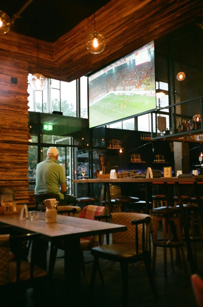 A man sitting in a cozy bar watching a sports game on a large screen, surrounded by wooden decor.