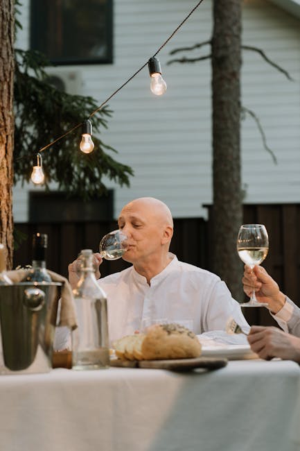 A bald elderly man sits at an outdoor dinner, enjoying a glass of white wine with ambient lighting.