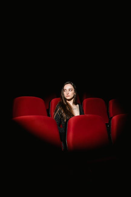 A woman sits alone in a dimly lit theatre with vibrant red seats, highlighting solitude.
