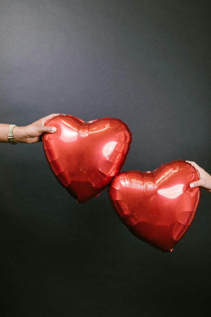 Two hands holding red heart-shaped balloons against a dark background symbolizing love and romance.